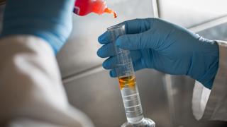 Close up image of a vial of clear liquid, two gloved hands can be seen holding the vial and adding drops of an orange  liquid into the vial.